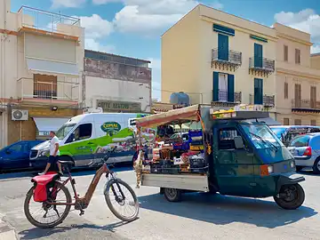 Ein Mietfahrrad von Sicula Bike steht mitten auf der Strasse neben der Ape eines Gemüsehändlers.