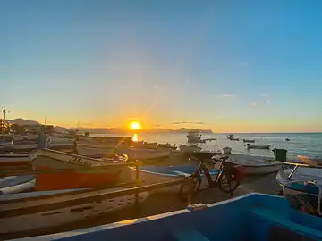Ein Fahrrad steht im Sonnenuntergang vor bunten Fischerbooten die am Strand von Aspra liegen.